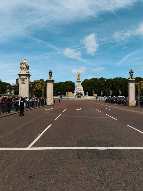 A wide view of a large, open paved area leading to a decorative monument with a golden statue on top, flanked by four ornate stone pillars topped with statues, with crowd barriers and people on either